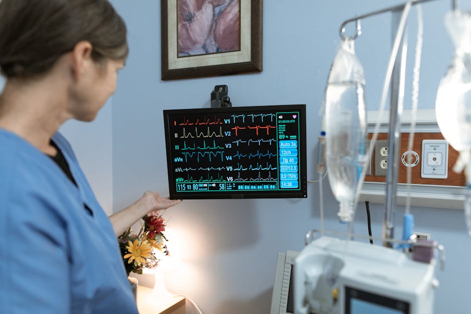 A nurse in blue scrubs examines a medical monitor displaying vital signs in a hospital setting.