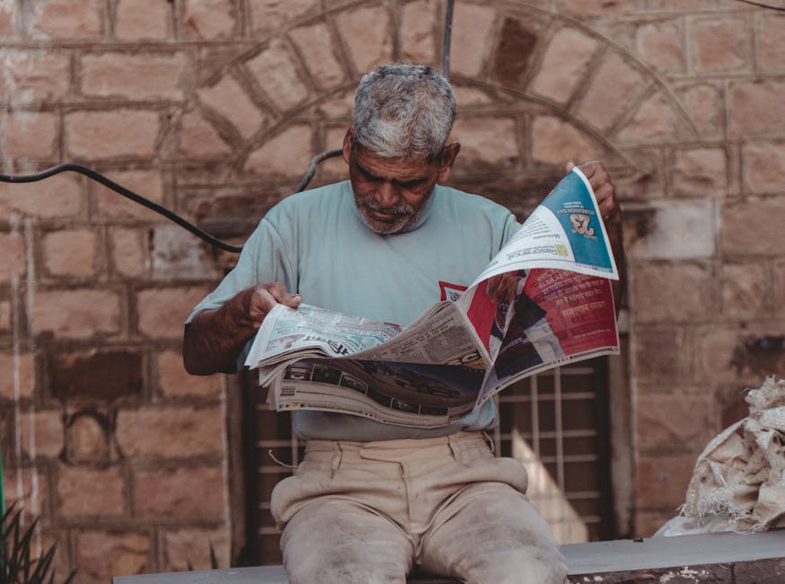 Senior man with gray hair sits outside, reading a newspaper in Mumbai, India.